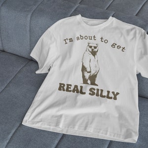 May include: White t-shirt with a brown bear wearing sunglasses and the text "I'm about to get real silly".