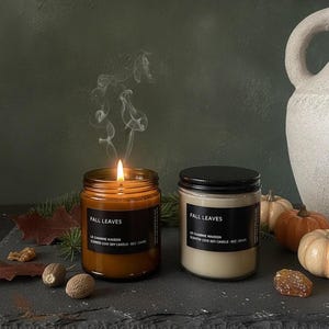 May include: Two glass jar candles labelled Fall Leaves by Le Charme Maison. One candle is amber-coloured with a lit wick producing smoke, while the other is cream-coloured with a black lid. Both are 8 oz soy candles.