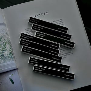 May include: An open book with the word "NATURE" printed on the top. Several black rectangular perfume boxes are arranged on the pages. Each box has white text indicating the fragrance name and "EAU DE PARFUM".