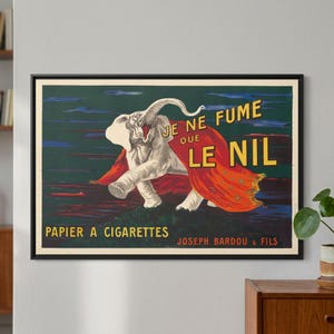 May include: Vintage framed advertisement featuring a white elephant wearing a red cape. The text reads "JE NE FUME QUE LE NIL" and "PAPIER A CIGARETTES JOSEPH BARDOU & FILS". The artwork has a dark green and blue background.