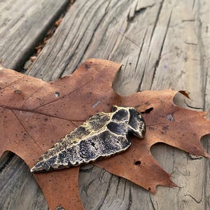 Könnte beinhalten: Ein goldener und schwarzer Anhänger in Pfeilspitzenform, der auf einem braunen Blatt liegt. Der Anhänger hat eine strukturierte Oberfläche und eine spitze Spitze.