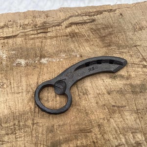 Puede incluir: Un abridor de botellas de metal negro con forma de herradura con un pequeño agujero en el centro. El abridor está sobre una superficie de madera.