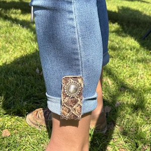 May include: Blue denim jeans with a decorative cuff featuring a brown, textured leather patch and a silver-toned metal embellishment. The jeans are cuffed at the ankle, showcasing the unique design element.