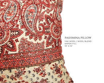 May include: A pashmina pillow featuring a red and cream paisley pattern with floral accents. Text on the pillow reads: PASHMINA PILLOW, YAK WOOL | WOOL BLEND, KAPOK FILL, 48.26 cm x 38.1 cm. The fabric has a woven texture with intricate red and black details.