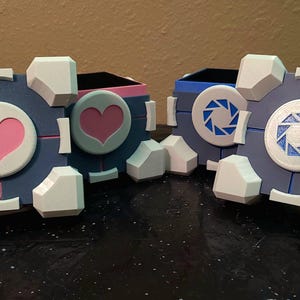 May include: Two blue and white cube-shaped storage boxes with a video game theme. One features a pink heart, the other a blue and white aperture symbol. The boxes have white corner accents and are likely for storing small items.