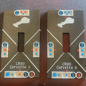 May include: Two black and white game pieces with the text "CR90 Corvette A" and "CR90 Corvette B". Each piece has a white ship silhouette, a white rectangle, and several red and blue circles and diamonds.