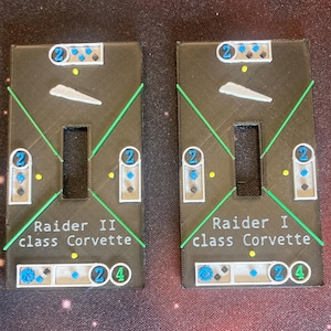 Könnte beinhalten: Zwei schwarze und weiße bedruckte Pappspielsteine mit grünen Linien und blauen und weißen Kreisen. Der Text auf den Steinen lautet "Raider II class Corvette" und "Raider I class Corvette".