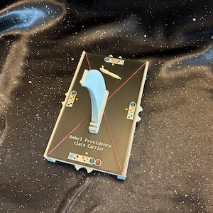 May include: A rectangular game board piece for a Rebel Providence class carrier, featuring a light blue plastic stand in the center. The surface is black with red diagonal lines, white text, and small circular icons with numbers and colored dots.