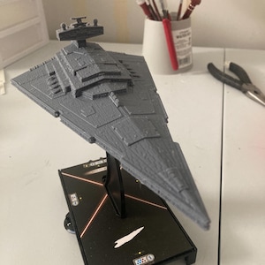 Puede incluir: Un modelo de nave espacial Star Destroyer gris sobre una base negra con un diseño de campo de estrellas. La base tiene un dial con números y un pequeño icono de nave blanca.