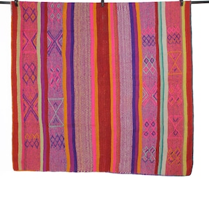 Könnte beinhalten: Ein farbenfrohes gewebtes Textil mit vertikalen Streifen in Rot-, Rosa-, Lila- und Orangetönen. Das Textil weist geometrische Muster in Rosa und Orange auf. Es hängt an einer schwarzen Stange mit Wäscheklammern.