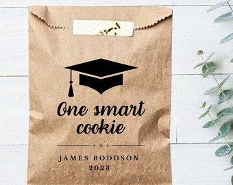 Könnte beinhalten: Eine braune Papiertüte mit einer schwarzen Graduation Cap und dem Text "One smart cookie" darauf gedruckt. Die Tüte trägt auch den Namen "JAMES BODDSON" und das Jahr "2023" aufgedruckt.