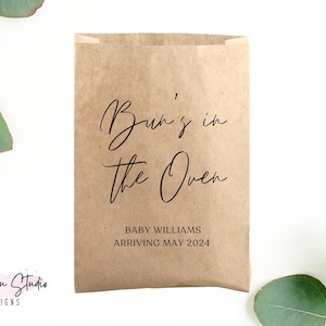 May include: A brown paper bag with the text "Bun's in the Oven" in black script. Below the text is "Baby Williams Arriving May 2024".