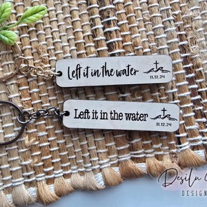 Op de afbeelding: Twee houten sleutelhangers met de tekst "Left it in the water 11.12.24" erop. De sleutelhangers zijn bevestigd aan metalen sleutelringen.