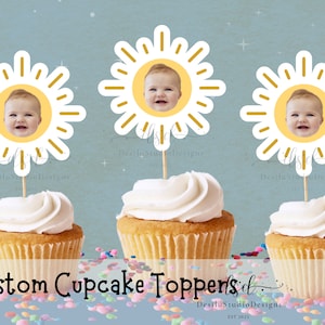 May include: Three yellow sun cupcake toppers with a white outline and a baby's face in the center. The toppers are on top of cupcakes with white frosting.
