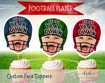May include: Three cupcake toppers featuring a baby's face in a football helmet. The helmets are blue, green, and red. Each helmet has the text "First Year Down" on it.