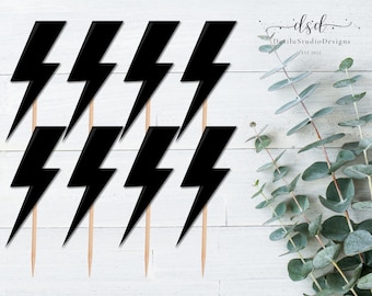May include: A collection of black lightning bolt cake toppers on wooden sticks. The toppers are arranged in a group, perfect for decorating a cake. The background is a white wooden surface with green leaves.