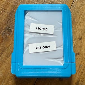 May include: A bright blue rectangular case with a clear plastic window. Two white labels inside read "LECTRIC" and "XP4 ONLY". The case is sitting on a wooden surface.
