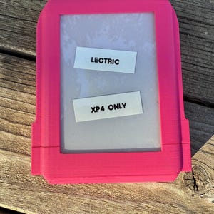 May include: A bright pink rectangular frame with the text "LECTRIC" and "XP4 ONLY" on white labels. The frame has a clear, transparent center, and is set against a wooden surface.