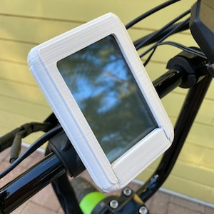 May include: A white rectangular display screen mounted on a black bicycle handlebar. The screen is encased in a white, textured frame. The background shows a glimpse of a yellow wall and a green and black bicycle component.