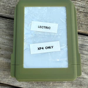 May include: An olive green rectangular case with a clear interior. Two white labels inside read "LECTRIC" and "XP4 ONLY". The case is on a weathered wooden surface.