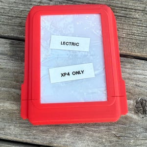 May include: A bright red rectangular case with a clear window. Two white labels inside the window read "LECTRIC" and "XP4 ONLY". The case is on a weathered wooden surface.
