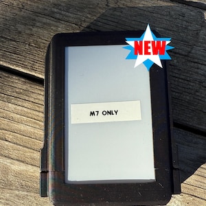 Könnte beinhalten: Ein schwarzes rechteckiges Gerät mit weißem Bildschirm und einem Aufkleber mit der Aufschrift "M7 ONLY". Eine blaue und rote Sternburst-Grafik mit dem Wort "NEW" befindet sich in der oberen rechten Ecke. Das Gerät liegt auf einer Holzoberfläche.