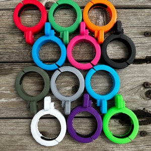 Puede incluir: Doce abrazaderas de plástico de diferentes colores, incluyendo rojo, verde, azul, rosa, gris, negro, blanco, morado y verde lima. Las abrazaderas están dispuestas en un círculo sobre una superficie de madera.