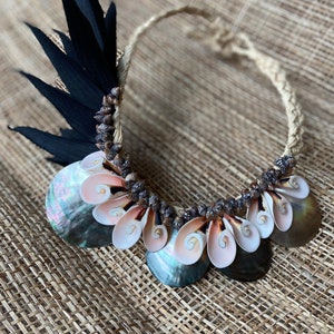 May include: A necklace made of natural materials, featuring a woven cord, black feathers, and a variety of seashells. The shells are arranged in a circular pattern, with the larger shells in the center and the smaller shells around the edges.