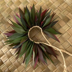 May include: A green and burgundy leaf lei with a natural fibre cord. The lei is made of many leaves that are woven together to form a circle.
