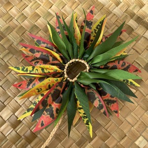 May include: A colourful lei made of green, yellow, red and black leaves. The lei is tied together with a brown cord and has a ring in the centre.