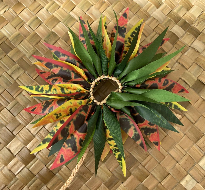 May include: A colourful lei made of green, yellow, red and black leaves. The lei is tied together with a brown cord and has a ring in the centre.