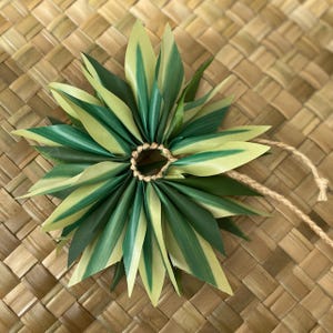May include: A green and white leaf lei with a brown string tied around the centre. The lei is on a woven brown background.