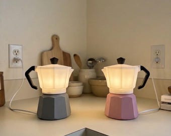 Puede incluir: Dos lámparas eléctricas con forma de cafetera moka, una con base gris y otra con base rosa. Ambas tienen una parte superior blanca, translúcida y facetada que se ilumina desde el interior, un asa negra y un pomo negro en la tapa.