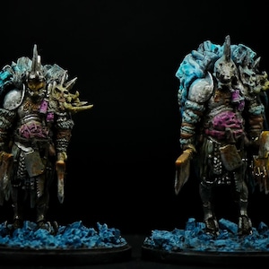 Puede incluir: Dos figuras en miniatura pintadas de guerreros armados con cuernos y espadas. Las figuras están en posición de pie sobre una base azul y gris. Las figuras están pintadas en una variedad de colores, incluyendo plata, oro, azul y rosa.