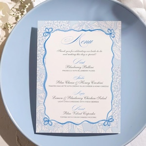 Op de afbeelding: Een lichtblauwe plaat met een menukaart met een bloemenontwerp en het woord "Menu" bovenaan. Het menu bevat gerechten zoals Blueberry Bellini en Blue Velvet Cupcake. Een gouden lepel en witte bloemen staan op de achtergrond.