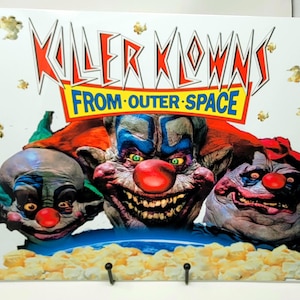 May include: A poster featuring three menacing clown faces with red noses and sinister smiles. The title "KILLER KLOWNS FROM OUTER SPACE" is displayed in bold, stylized lettering. Popcorn accents the bottom and background.