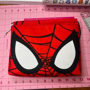 May include: A red drawstring bag featuring the face of Spider-Man. The bag has a black web design and large white eye shapes. The bag is rectangular and appears to be made of fabric.