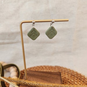 Könnte beinhalten: Ein Paar grüne, blattartige Ohrringe auf einem goldenen Ständer. Die Ohrringe sind aus Polymer Clay gefertigt und haben eine quadratische Form.