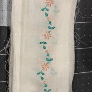 May include: A strip of off-white fabric featuring a vertical embroidered vine pattern. The design consists of small, light peach-colored cross-shaped flowers connected by a winding green stem with tiny leaves, creating a continuous floral motif.