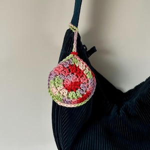 Puede incluir: Un bolso de pana azul oscuro con un amuleto de flor tejido a crochet en tonos de rosa, rojo, verde y blanco.