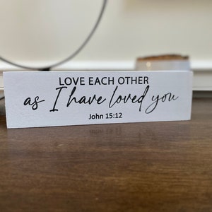May include: White wooden sign with black text that reads "LOVE EACH OTHER as I HAVE LOVED YOU" and "John 15:12".