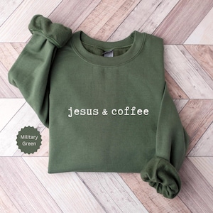 May include: A military green sweatshirt with the text "jesus & coffee" in white. The sweatshirt is folded on a light brown wooden surface. The sleeves are cuffed. A small label in the lower left corner indicates the color is military green.