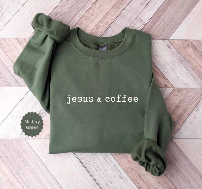 May include: A military green sweatshirt with the text "jesus & coffee" in white. The sweatshirt is folded on a light brown wooden surface. The sleeves are cuffed. A small label in the lower left corner indicates the color is military green.
