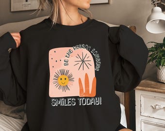 May include: Black sweatshirt with a graphic design. The design features a smiling sun, a starburst, and the text "Be the reason someone smiles today!" in white. The design is on a peach-colored background.