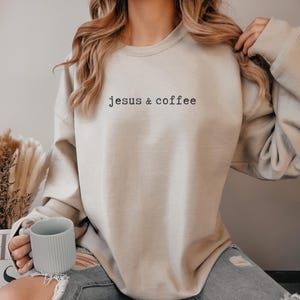 May include: Beige sweatshirt with the text "jesus & coffee" in black. The sweatshirt is a neutral color and has a relaxed fit. A person is holding a light gray mug.