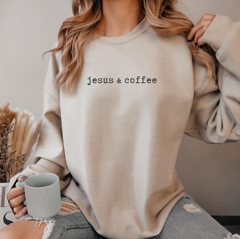 May include: Beige sweatshirt with the text "jesus & coffee" in black. The sweatshirt is a neutral color and has a relaxed fit. A person is holding a light gray mug.