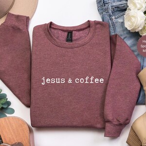 May include: Maroon crewneck sweatshirt with the text "jesus & coffee" in white. The sweatshirt is made of a soft material and has long sleeves. The image also includes a pair of jeans, a pair of boots, and a guitar.