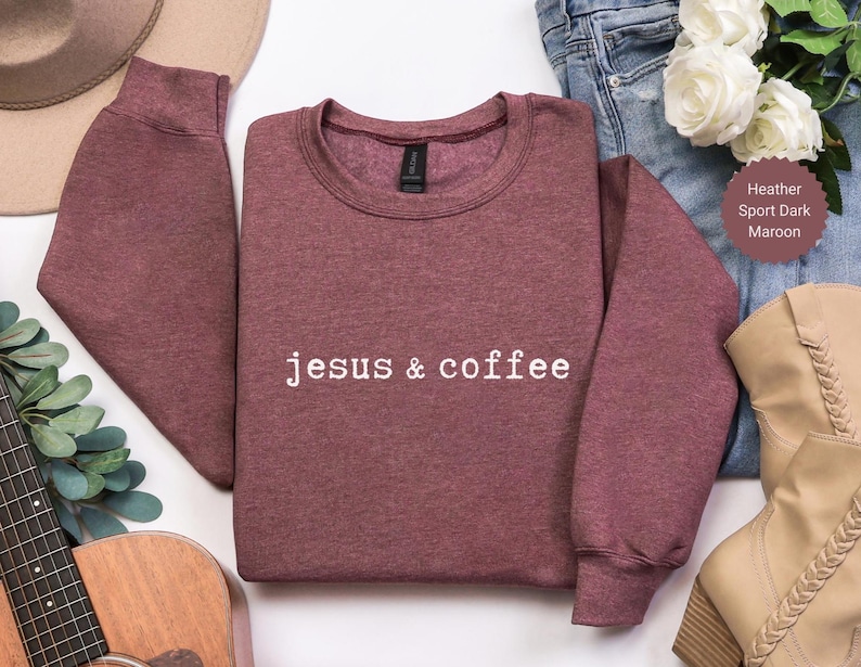 May include: Maroon crewneck sweatshirt with the text "jesus & coffee" in white. The sweatshirt is made of a soft material and has long sleeves. The image also includes a pair of jeans, a pair of boots, and a guitar.