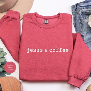 May include: A red heather sports scarlet sweatshirt with the text "jesus & coffee" in white. The sweatshirt is displayed with a guitar, jeans, a hat, and flowers.