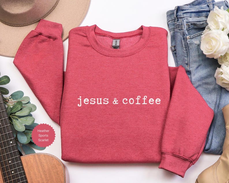 May include: A red heather sports scarlet sweatshirt with the text "jesus & coffee" in white. The sweatshirt is displayed with a guitar, jeans, a hat, and flowers.